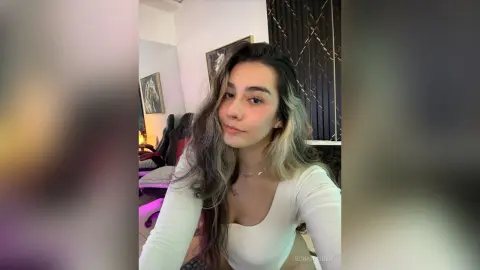 BonnieRider poses in a white long-sleeve top with her long, wavy hair styled over one shoulder in a room with a desk chair and wall art.