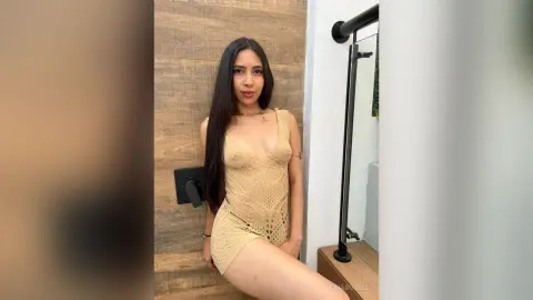 IrinaRussel poses in a tan lace bodysuit, showing her long dark hair and cleavage while sitting against a wooden wall.