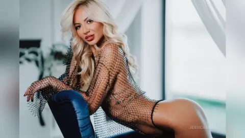 Blonde woman in a sheer black mesh top and blue jeans posing in a bright, airy room.