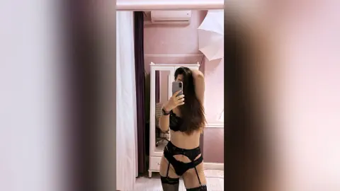 Young woman in black lace lingerie and stockings posing for a mirror selfie in a bright bedroom.