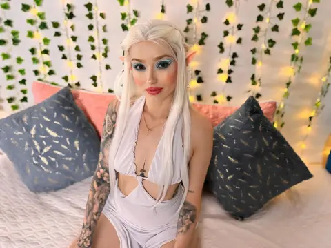 Woman in white fantasy cosplay with long blonde hair sitting on a bed with decorative pillows.