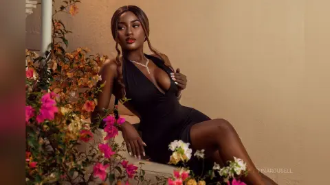 TinaRousell wearing a low-cut black dress, posing outdoors next to bright pink and white flowers.