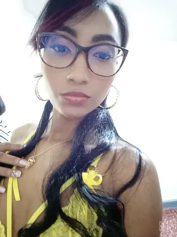 Young woman with long dark hair and glasses wearing a yellow lace top in a bright indoor setting.