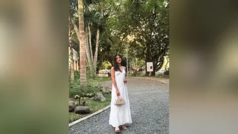 Elegant woman in a white summer dress standing on a gravel path in a lush green garden setting.