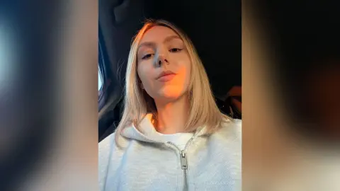 Blonde woman in a white zip-up hoodie sitting in a car with warm sunset lighting.