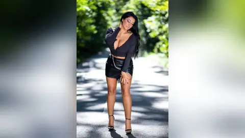 MissyJolie stands on a paved path wearing a black long-sleeve crop top and a black leather mini skirt with black high heels.