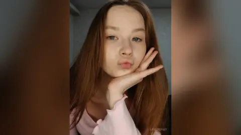 Young woman with long brown hair wearing a pink long-sleeve shirt making a playful pouty face indoors.