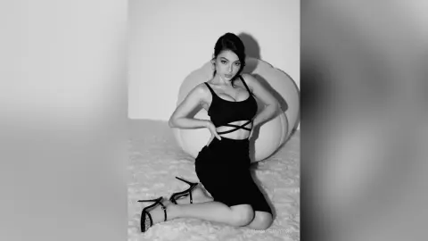 Elegant woman in a black crop top and skirt sitting on a white rug in a moody black and white studio portrait.