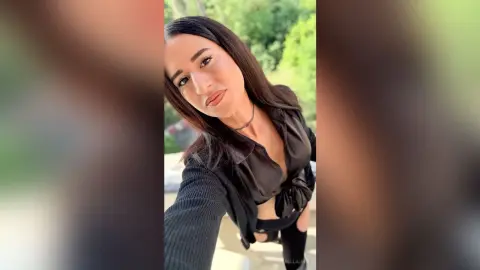 BellaLeen poses outdoors with long dark hair, wearing a black button-down shirt tied at the waist and black pants.