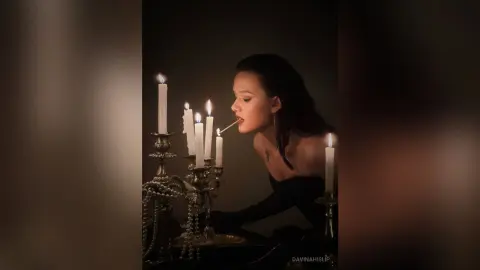 Moody portrait of a woman in a black dress biting a lit candle in a dark, atmospheric setting.