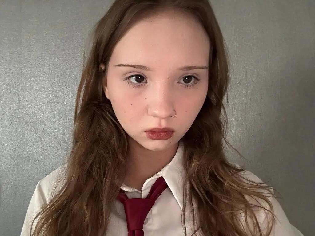 Young woman with long brown hair wearing a white shirt and red tie against a grey wall background.