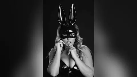 Glamorous woman in a black latex bunny mask and corset posing in a moody black and white studio portrait.