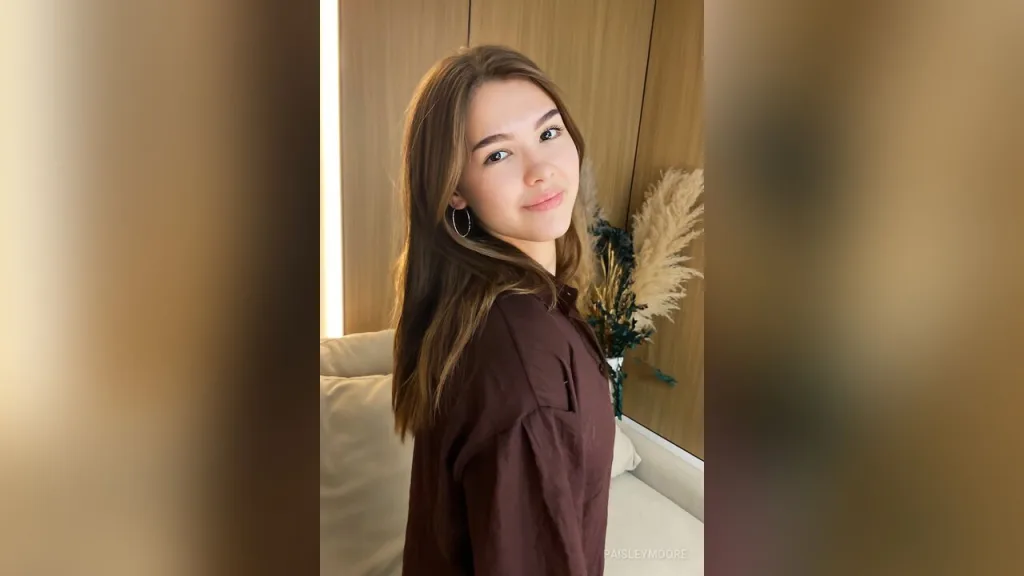 PaisleyMoore — PaisleyMoore smiles at the camera while wearing a brown long-sleeve shirt in a living room setting.