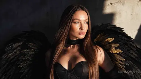 KatyMonro poses with large black and gold feathered wings, wearing a black corset and a black choker, with her long brown hair down.