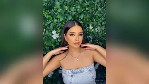 Beautiful woman with glamorous makeup in a light denim tube top posing against a lush green leaf wall.
