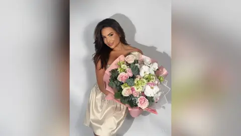 Smiling woman in a champagne satin dress holding a large bouquet of pink and white flowers against a white wall.