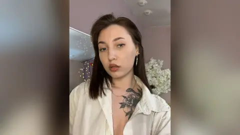 Young woman with long brown hair wearing a white button-down shirt and gold earrings in a soft-lit room.