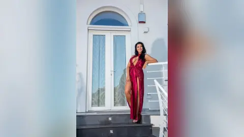MissyJolie stands on a balcony wearing a long, red lace slit dress, leaning against a white railing.
