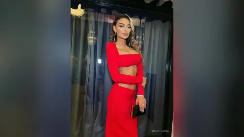 Anastasia poses in a red long-sleeved crop top and matching skirt, standing in front of dark blue curtains.