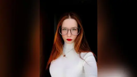 Redheaded woman in a white turtleneck and glasses posing with a moody, elegant aesthetic.