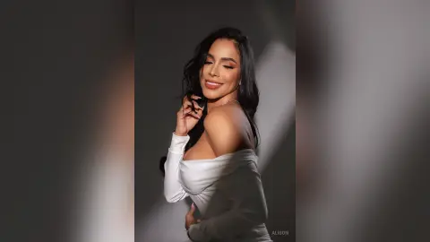 Alison smiles while posing with one hand near her face, wearing a white off-the-shoulder long-sleeve top against a dark grey background.