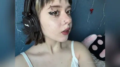Young woman with dark eyeliner and headphones in a cozy bedroom setting with a relaxed mood.