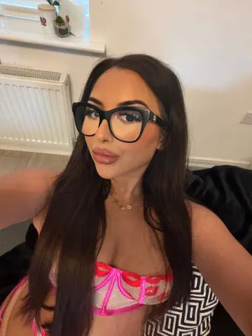 Brunette woman with black glasses and pink lace lingerie posing for a selfie in a bright indoor setting.