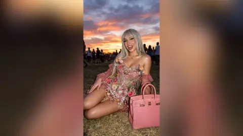 Smiling blonde woman in a pink floral mini dress sitting on grass during a dreamy sunset outdoor event.