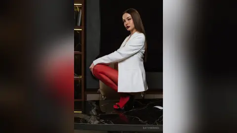 Elegant woman in a white blazer and red tights sitting on a marble floor in a moody, sophisticated setting.