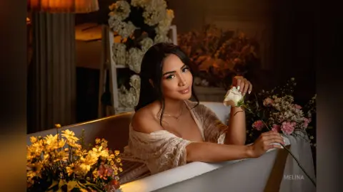 Elegant woman in a lace cream dress relaxing in a white bathtub surrounded by romantic floral arrangements.