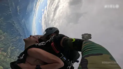 RossiNoleen soars through the sky in a helmet and black harness, wearing a green camouflage bodysuit against a backdrop of clouds and mountains.
