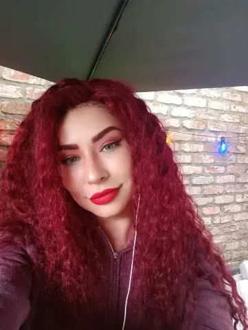 DanyelleVee smiles with long, curly red hair and red lipstick, wearing a purple zip-up top against a brick wall background.