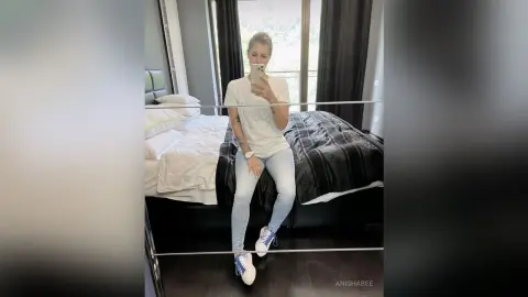 AnishaBee poses in a white t-shirt and light blue jeans while sitting on the edge of a bed in a bedroom.