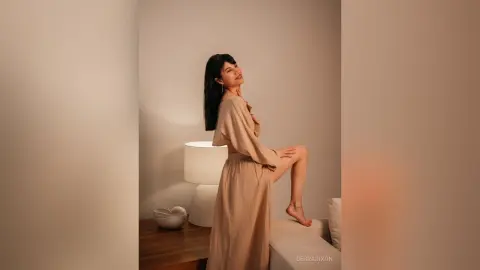 Smiling woman in a beige linen maxi dress relaxing in a warm, minimalist bedroom setting.
