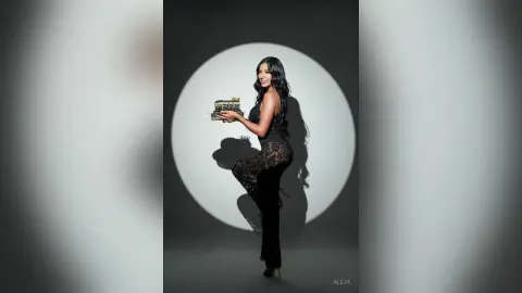 Aleja stands in profile wearing a black lace bodysuit while holding a tray of treats against a grey circular backdrop.