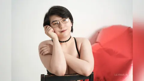 Smiling young woman with glasses in a black bikini top relaxing in a bright, minimalist indoor setting.