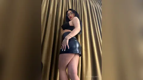 Confident woman in a black crop top and leather mini skirt posing against gold curtains.