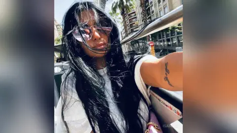 Young woman with long dark hair and sunglasses smiling in a sunny outdoor urban setting.