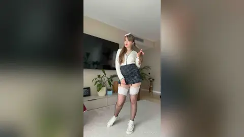 QuinnTakara poses in a white cardigan, plaid skirt, and white thigh-high stockings with black fishnets in a bright living room.