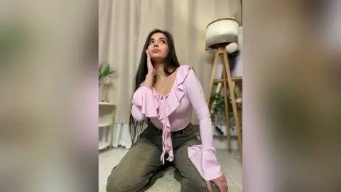 LettyKaminska poses with a hand to her face, wearing a long-sleeved pink ruffled bodysuit and grey jeans.