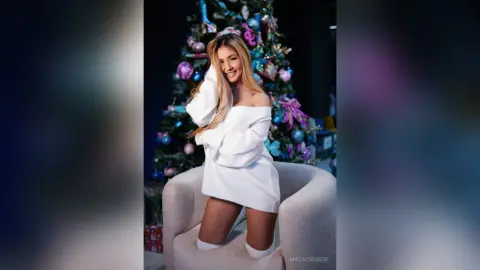 Anita smiles while wearing a white off-the-shoulder sweater, sitting in a light grey chair near a decorated Christmas tree.