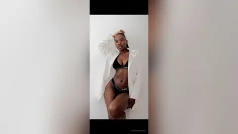 Confident Black woman in black lingerie and white oversized shirt posing against a minimalist white background.