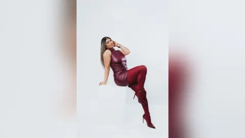 Elegant woman in a burgundy leather dress and matching boots posing on a white pedestal in a studio setting.
