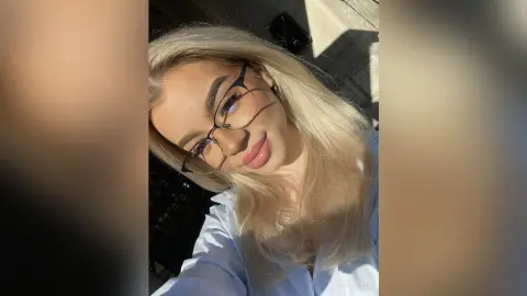 Smiling blonde woman in a light blue shirt wearing glasses in bright, warm sunlight.