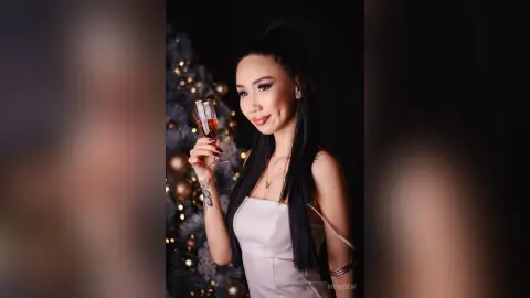 Elegant woman in a white dress holding a champagne glass near a festive Christmas tree in a moody setting.
