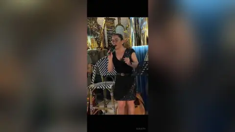 Elegant woman in a black cocktail dress speaking into a microphone at a glamorous indoor event.