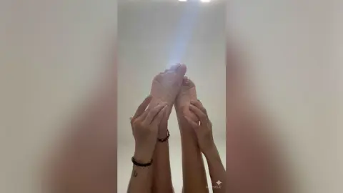 SevenEle holds her bare feet up high against a white background, showing off her soles and toes.