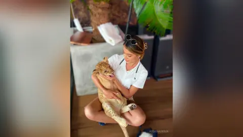 Smiling woman in a white shirt cuddling a small tan puppy on a sunny outdoor patio.