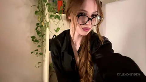 KylieHoneydew wearing black glasses and a black shirt, posing in front of a green plant.