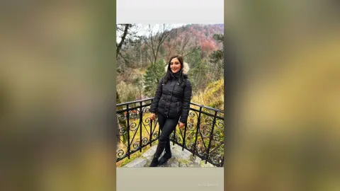 Smiling woman in a black puffer jacket and leggings standing on a balcony overlooking a scenic autumn forest.